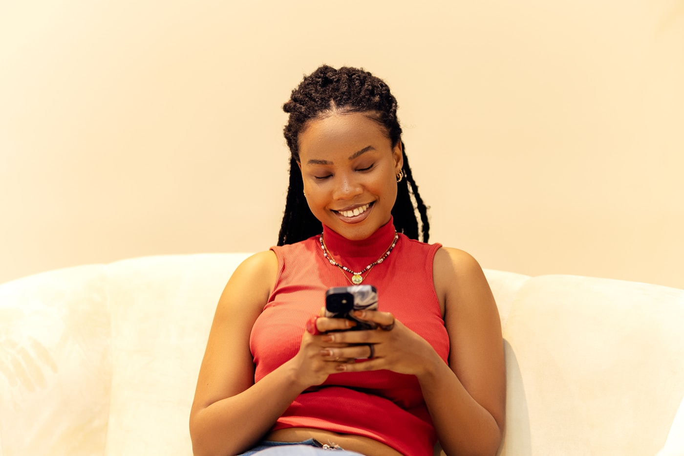 Mulher sorrindo enquanto olha para o celular, sentada em um sofá claro